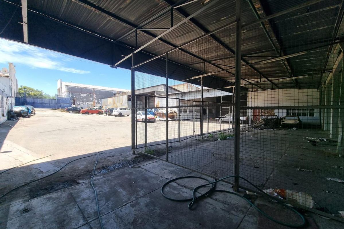 Industrial rear yard area featuring a large covered steel-framed structure with corrugated metal roofing and wire mesh partitioning over a concrete base.