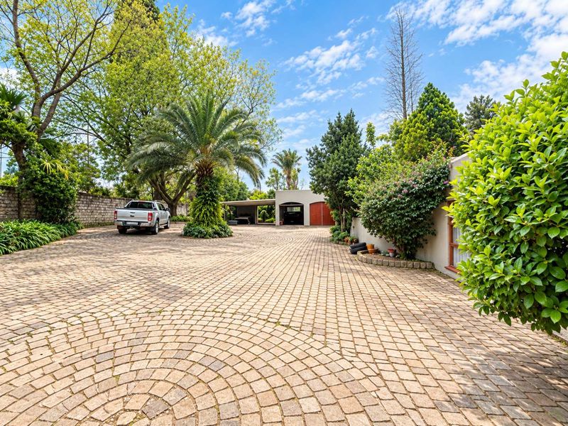 Spacious front exterior featuring a circular cobblestone driveway, mature landscaping, and a multi-car carport and garage structure.