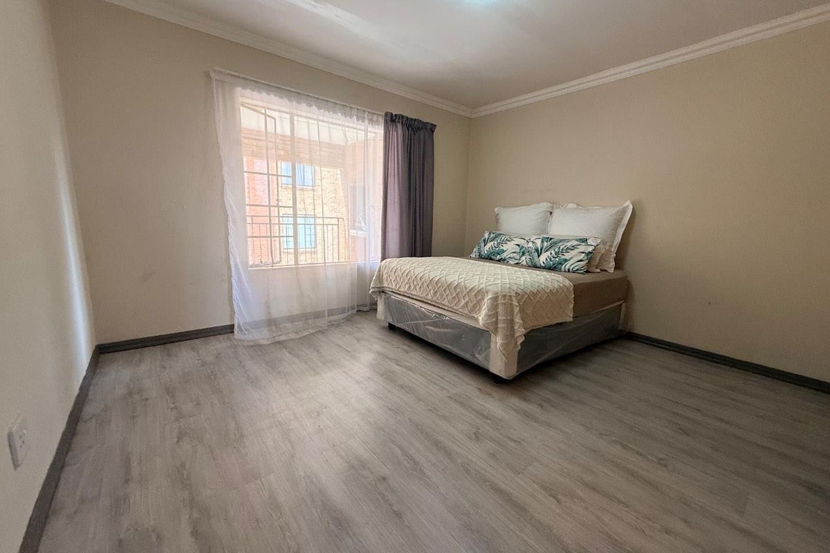 A spacious bedroom featuring modern light-toned laminate flooring, neutral wall finishes, and a large window with security bars allowing for natural light.