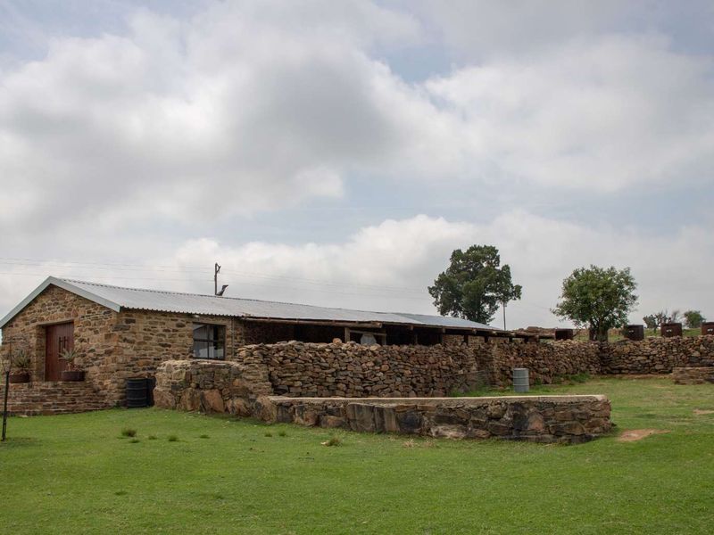 A rustic stone-built farm building featuring a corrugated metal roof, stone retaining walls, and expansive surrounding lawn area.