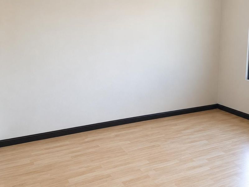 A bright, empty bedroom featuring light-toned laminate flooring, dark skirting boards, and neutral wall finishes.