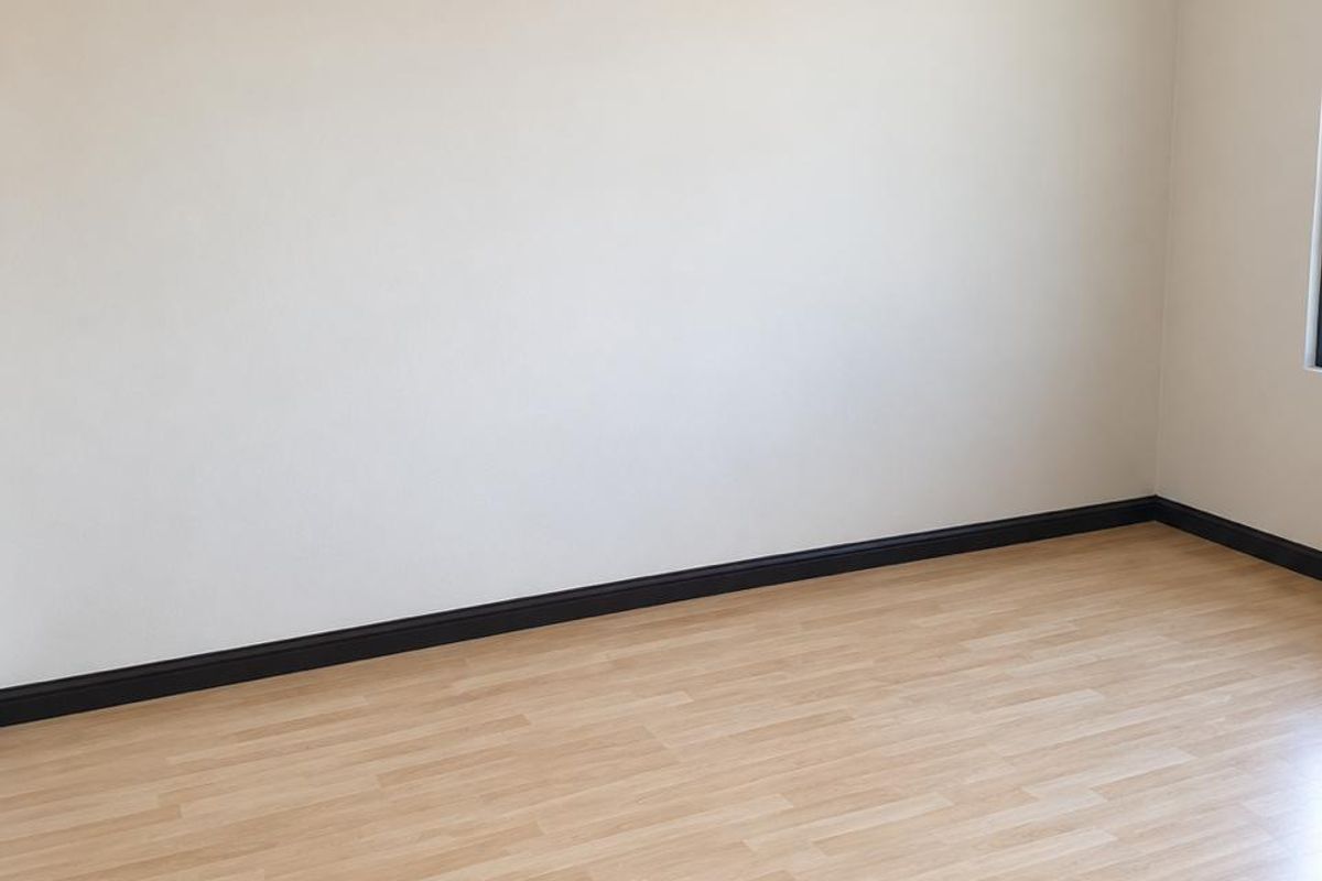 A bright, empty bedroom featuring light-toned laminate flooring, dark skirting boards, and neutral wall finishes.