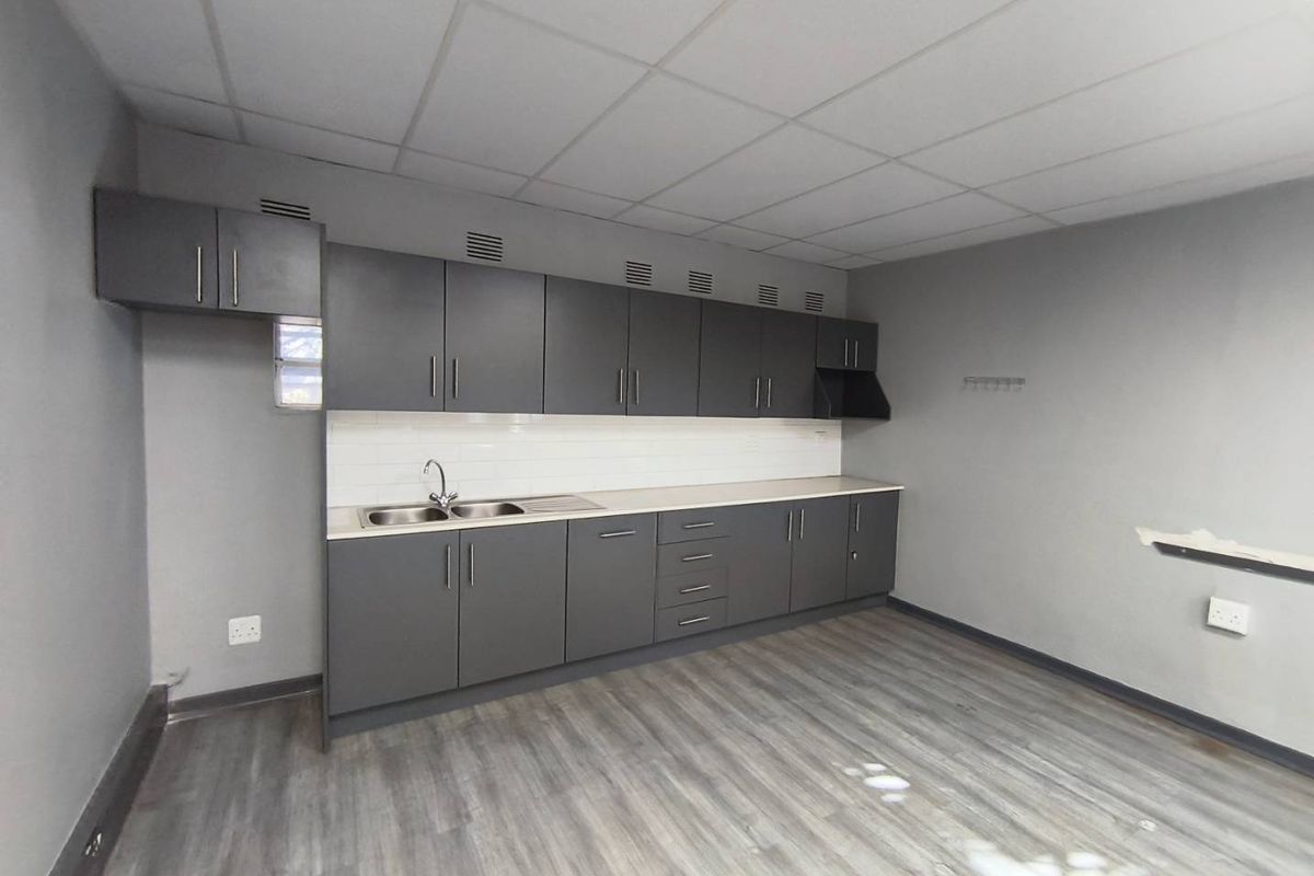 A functional kitchenette featuring dark grey cabinetry, a stainless steel sink, tiled backsplash, and durable laminate flooring in an open industrial office space.