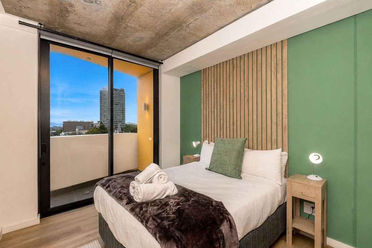 Modern bedroom featuring an exposed concrete ceiling, timber slat accent wall, and large sliding glass doors opening onto a private balcony.
