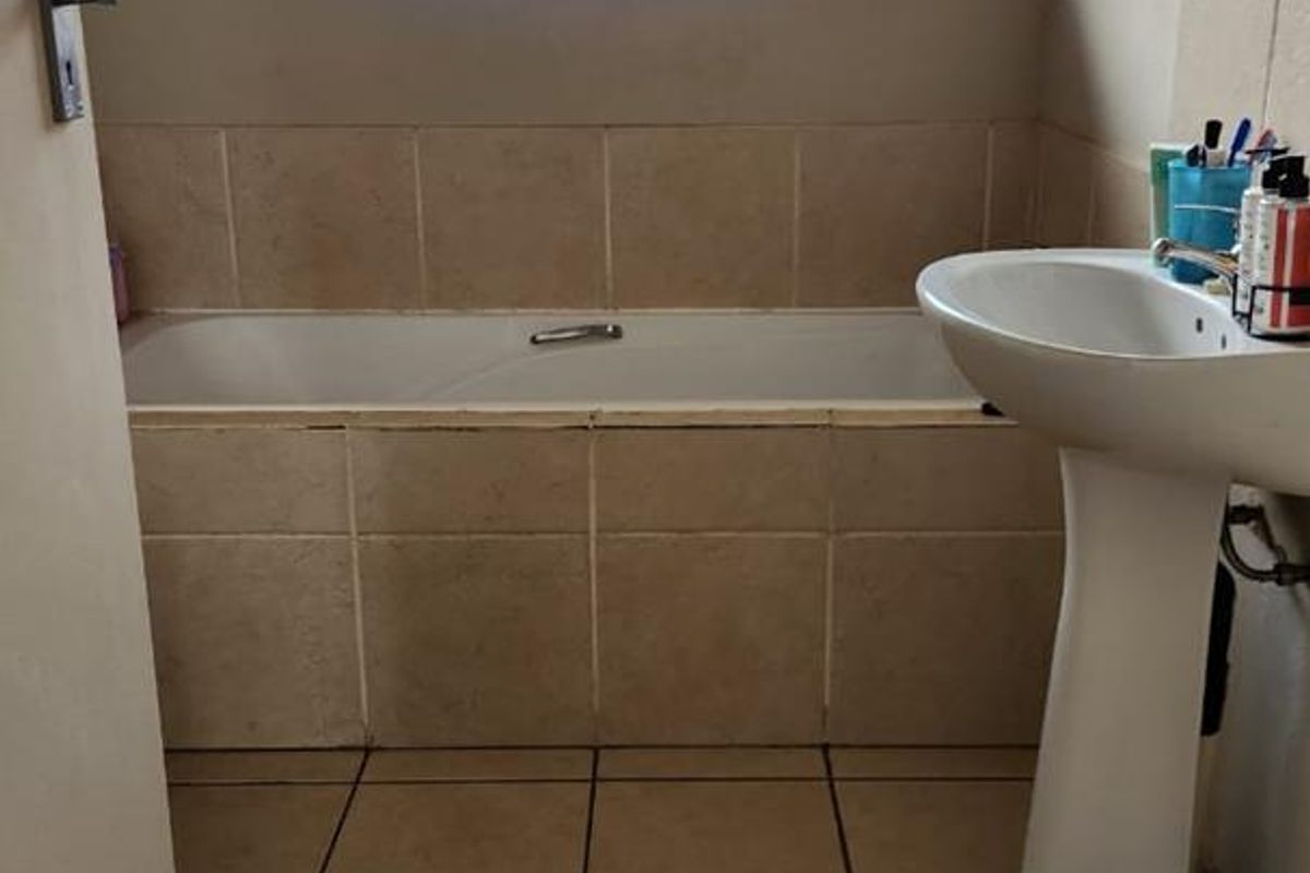 A functional bathroom featuring a tiled bathtub surround, pedestal basin, and ceramic floor tiles, illuminated by a window with fitted blinds.