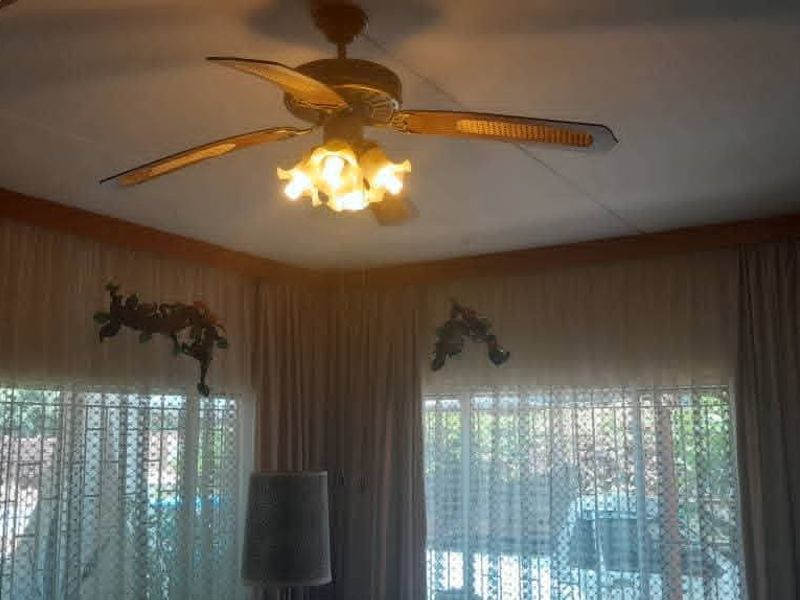 A living room area featuring a ceiling fan with integrated lighting and large windows fitted with security bars and curtain tracks.