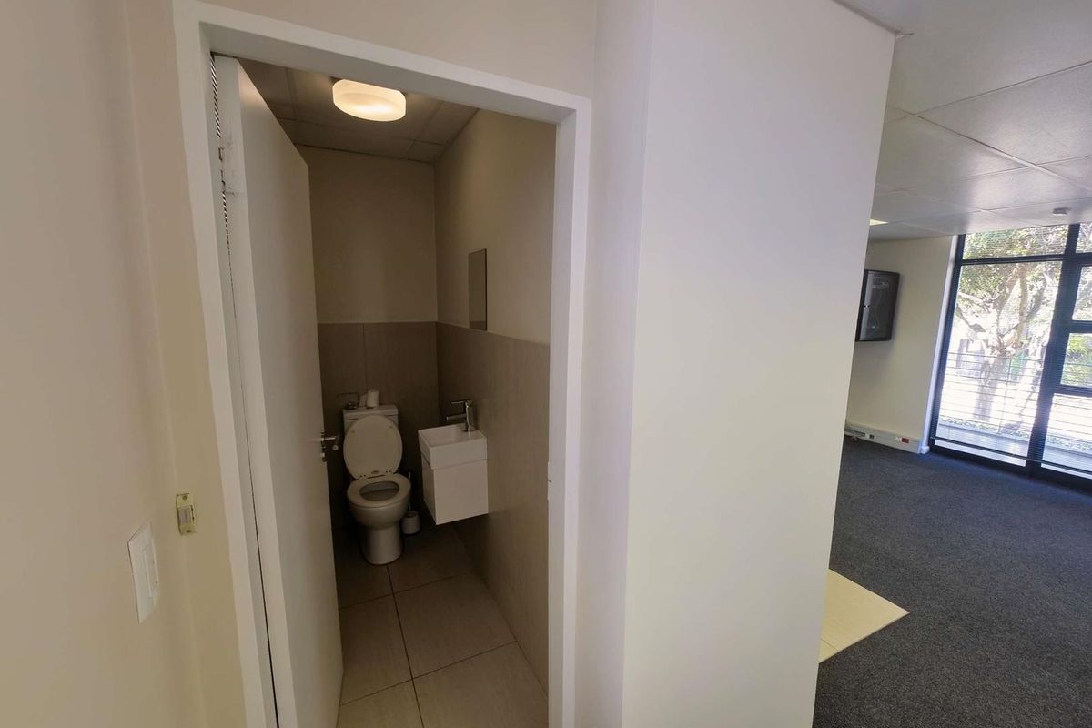 A compact office restroom featuring a wall-mounted basin and toilet, adjacent to an open-plan office area with large windows.