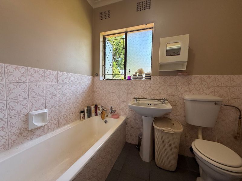 A dated bathroom featuring a standard bathtub, pedestal basin, and toilet with original wall tiling and security bars on the window.