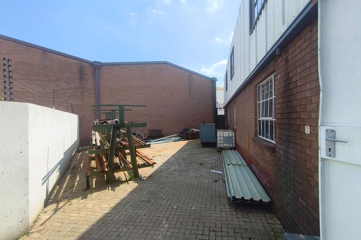 A paved rear industrial yard area featuring brick boundary walls, electric fencing, and steel-framed windows along the building exterior.