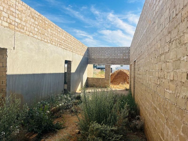 An unfinished residential construction site showing exposed brick and concrete walls with an open-air layout and overgrown ground.