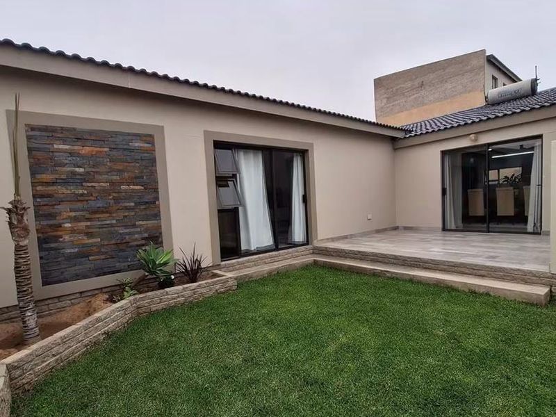A well-maintained rear exterior featuring a landscaped lawn, stone-clad feature wall, tiled patio area, and modern sliding glass doors providing indoor-outdoor flow.