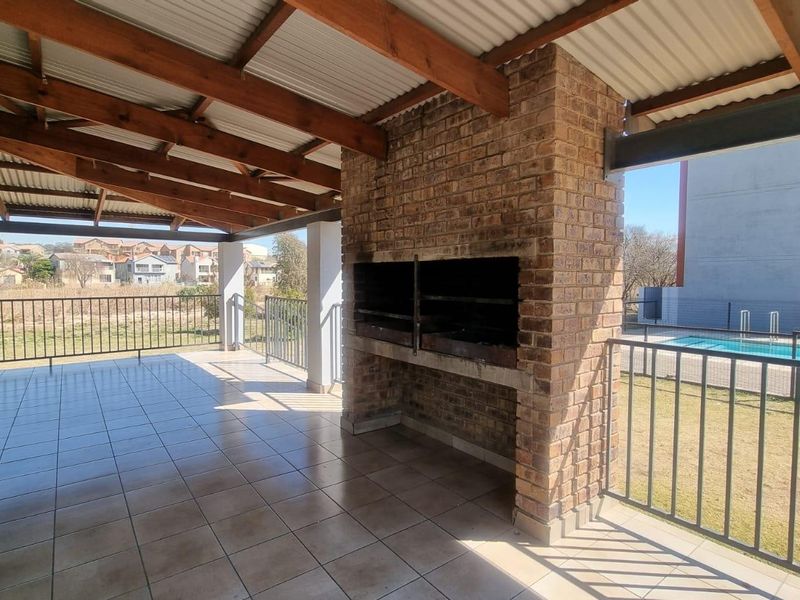 Covered patio area featuring a built-in brick braai, tiled flooring, and exposed timber rafters, overlooking the complex pool and garden area.