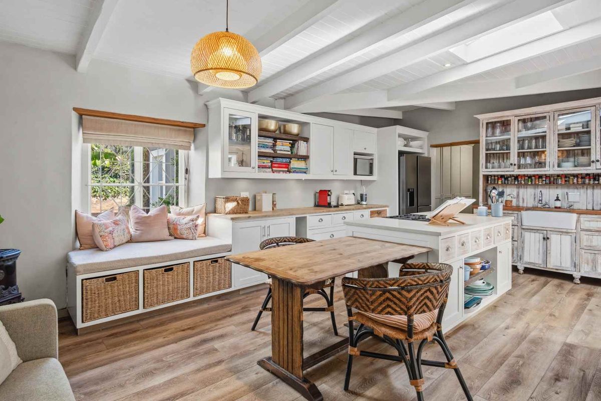 A bright, farmhouse-style kitchen featuring white cabinetry, a central island, a farmhouse sink, exposed beam ceilings, and a cozy built-in window seat.