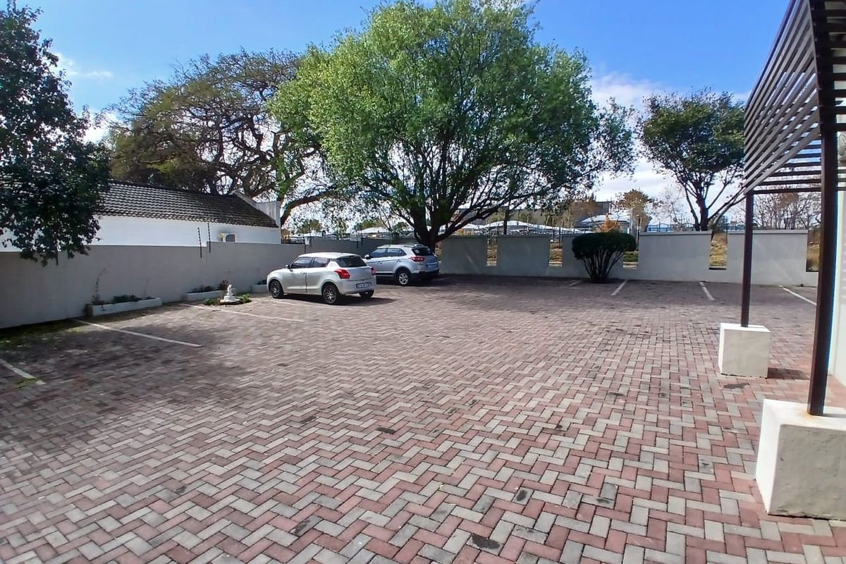 A paved outdoor parking area featuring a perimeter wall, mature trees, and a partial pergola structure.