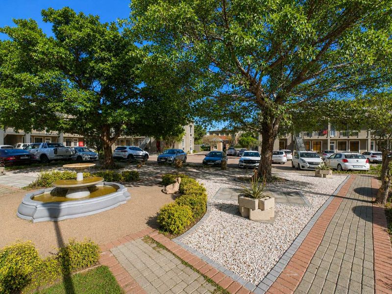 A well-maintained office complex courtyard featuring a central fountain, paved walkways, mature trees, and landscaped garden beds.