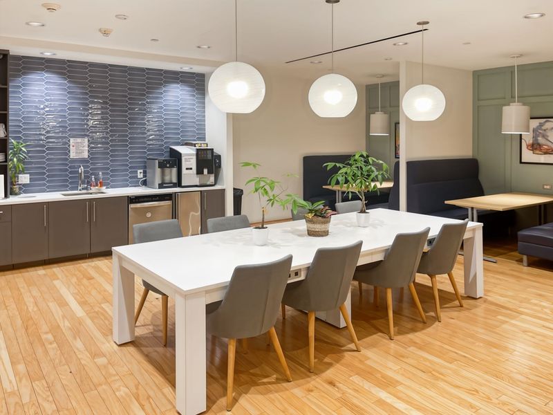 A modern open-plan office breakroom featuring a kitchenette with tiled backsplash, integrated sink, hardwood flooring, and comfortable built-in booth seating with pendant lighting.