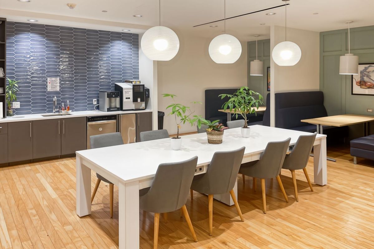 A modern open-plan office breakroom featuring a kitchenette with tiled backsplash, integrated sink, hardwood flooring, and comfortable built-in booth seating with pendant lighting.