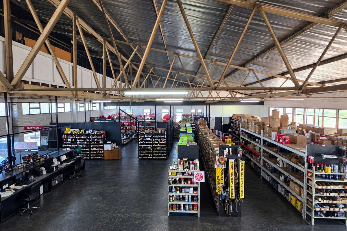 Spacious industrial warehouse featuring high exposed steel roof trusses, mezzanine level, and extensive fixed steel shelving units for storage and retail display.
