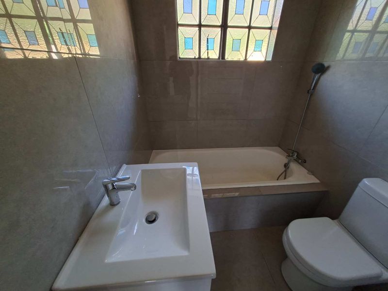 A clean, modern bathroom featuring a built-in bathtub, ceramic vanity basin, and toilet, finished with neutral wall tiles and a decorative leaded glass window.