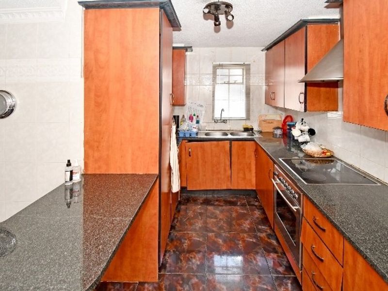 A functional kitchen featuring wood-finish cabinetry, granite countertops, built-in oven, electric hob, and a double sink with tiled finishes throughout.