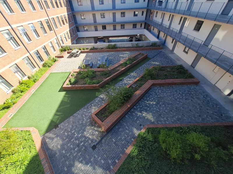 An elevated view of a communal courtyard featuring brick-paved pathways, raised garden planters, artificial grass, and surrounding apartment building walkways.