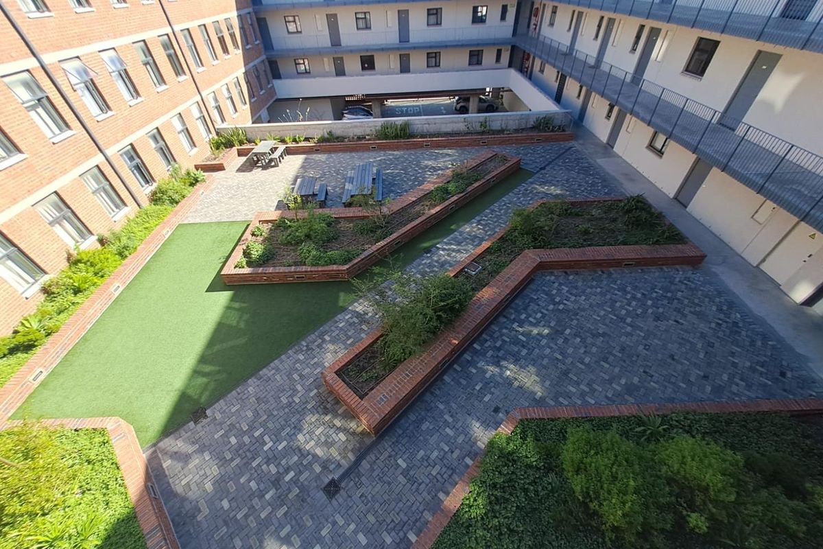 An elevated view of a communal courtyard featuring brick-paved pathways, raised garden planters, artificial grass, and surrounding apartment building walkways.