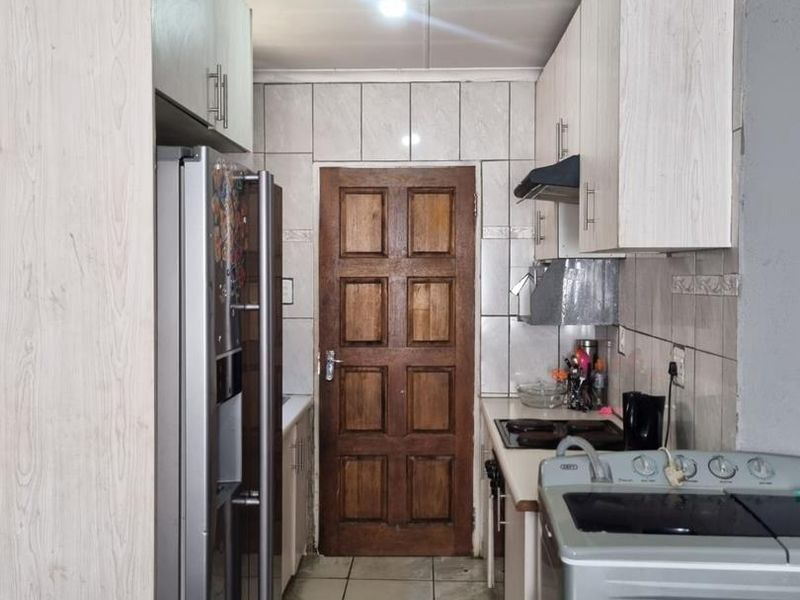 A compact kitchen featuring light-toned built-in cabinetry, tiled backsplash, and tiled flooring, leading to a solid wood door.