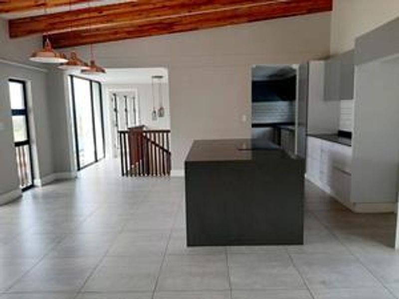 Spacious open-plan living and kitchen area featuring exposed timber beams, a central kitchen island, modern cabinetry, and tiled flooring with staircase access.