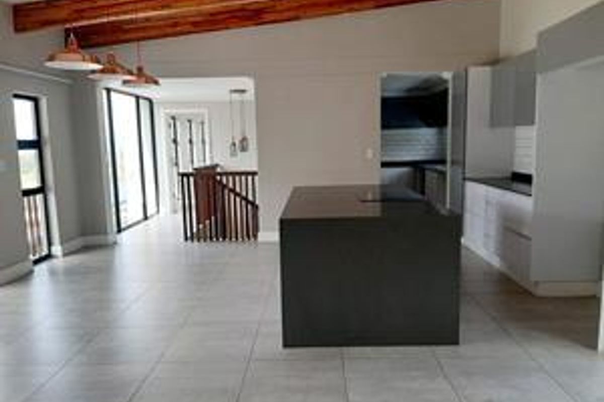 Spacious open-plan living and kitchen area featuring exposed timber beams, a central kitchen island, modern cabinetry, and tiled flooring with staircase access.