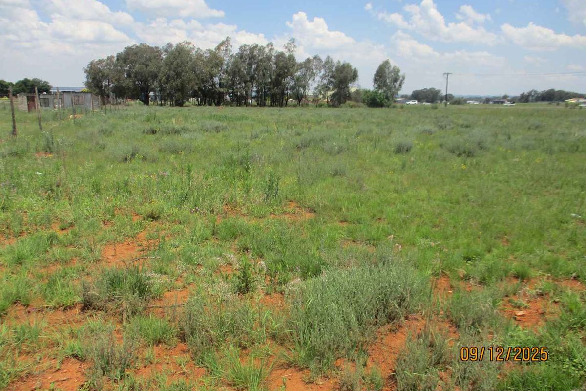 A vacant residential plot featuring natural grass cover, perimeter fencing, and mature trees in the background.