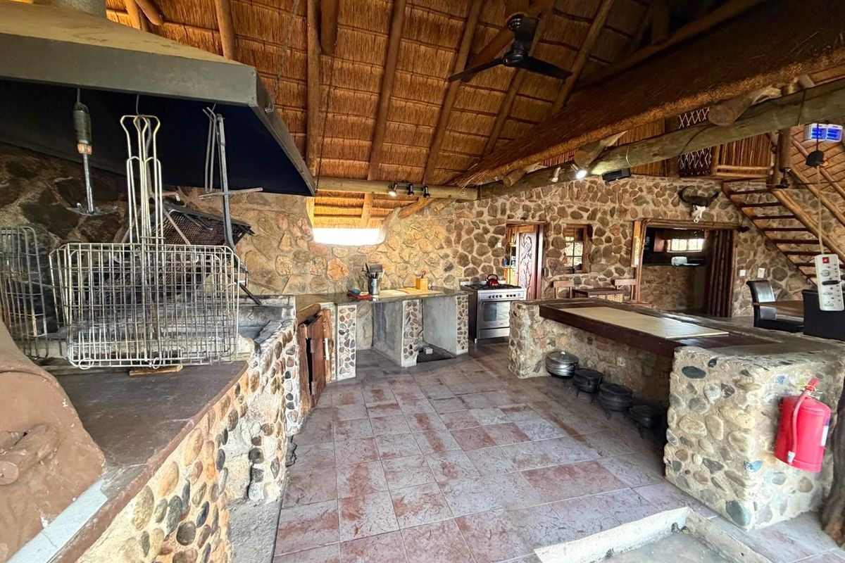 Rustic open-plan kitchen and entertainment area featuring stone masonry walls, a built-in braai, stone breakfast counter, and traditional thatch roofing.