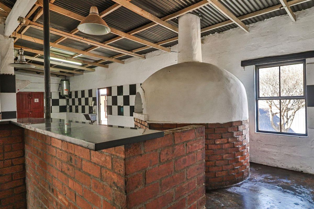 Rustic kitchen area featuring a large built-in brick pizza oven, granite-topped breakfast counter, and exposed timber roof trusses with industrial-style lighting.