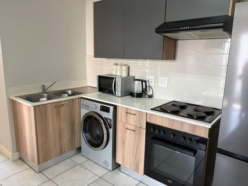 Modern kitchen featuring wood-finish cabinetry, contrasting grey upper cabinets, integrated electric hob, oven, extractor fan, and a stainless steel sink with tiled backsplash.
