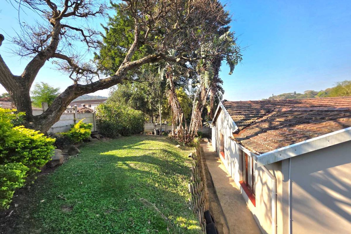 A view of the rear garden area featuring a lawn, mature trees, and the side elevation of the house with tiled roofing.