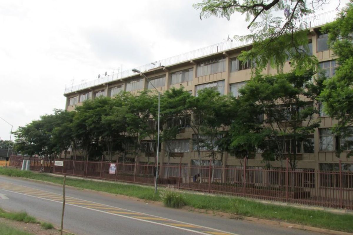 A street-level view of a multi-story apartment building featuring a fenced perimeter and mature trees along the roadside.