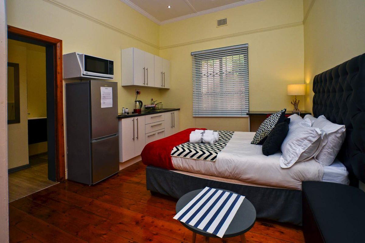 An open-plan studio apartment featuring a kitchenette with built-in cabinetry, timber flooring, and a bedroom area with neutral walls and classic cornicing.