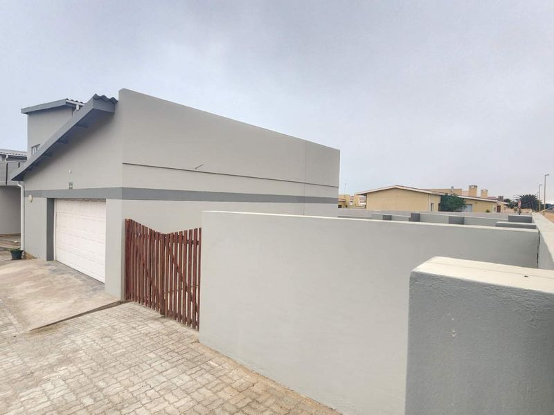 Exterior view of a modern townhouse featuring a paved driveway, a single garage, and a secure boundary wall with a timber gate.