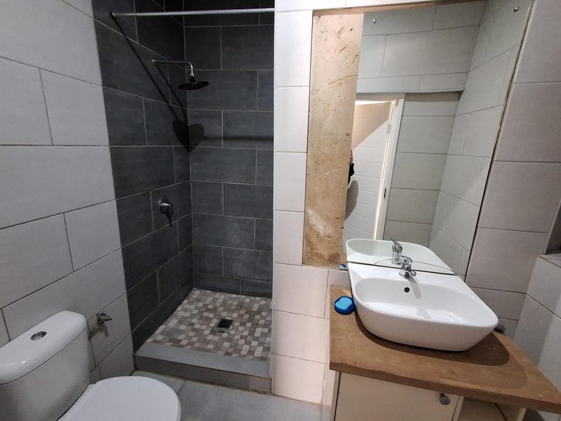 A functional bathroom featuring a walk-in shower with grey tiling, a modern vanity basin, and a toilet. The wall finish near the mirror is incomplete.