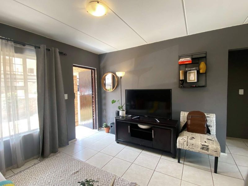 A living room featuring tiled flooring, neutral grey walls, and natural light from an adjacent entrance door and window.