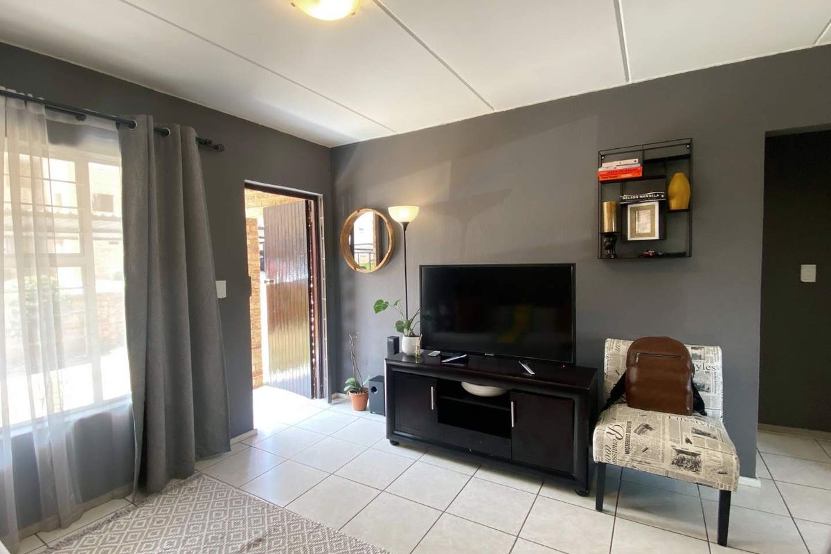 A living room featuring tiled flooring, neutral grey walls, and natural light from an adjacent entrance door and window.