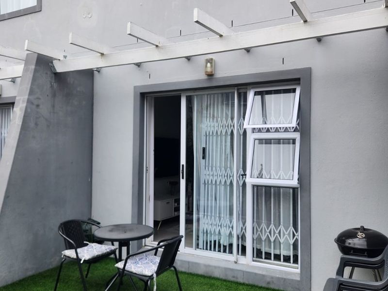 A compact ground-floor patio featuring artificial turf, paved sections, and a timber pergola, providing direct access to the interior via a sliding glass door.