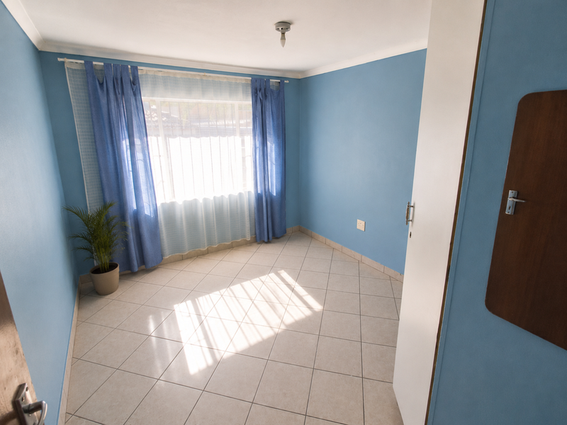 A bright, empty bedroom featuring light-colored floor tiles, blue painted walls, and a window with curtains.