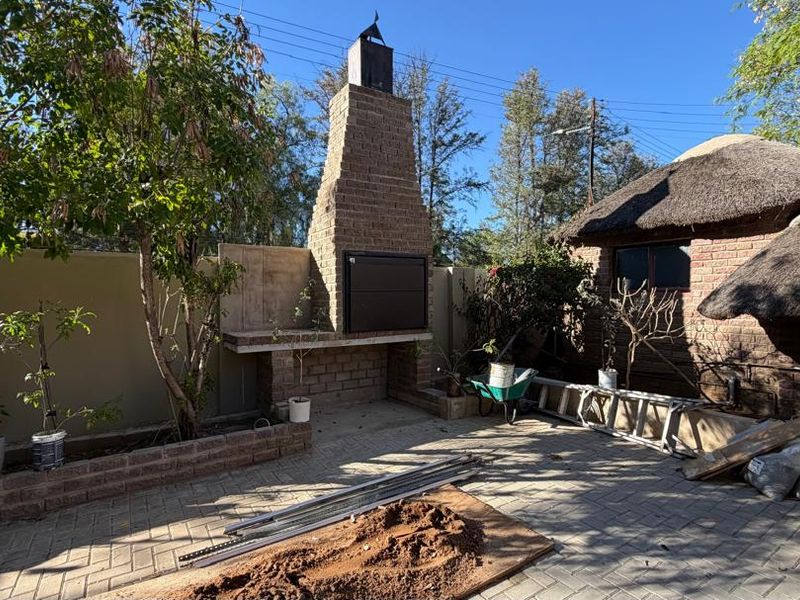 Rear outdoor area featuring a built-in brick braai, paved patio space, and traditional thatched roof structure, currently undergoing minor maintenance.