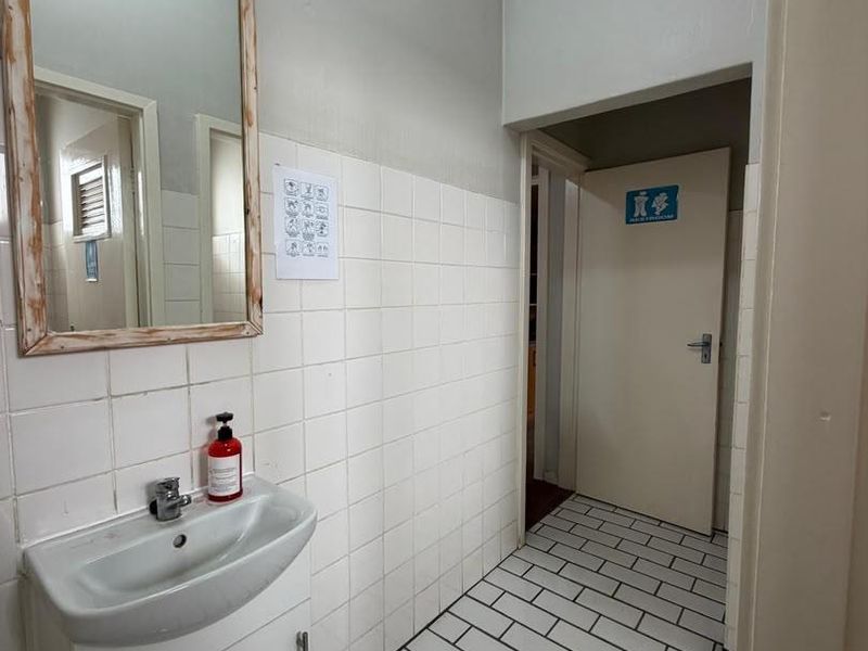 A functional office restroom area featuring a wall-mounted basin, tiled splashback, and tiled flooring, providing basic sanitary facilities for the workspace.