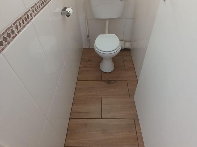 A compact guest bathroom featuring wood-look floor tiles, white ceramic wall tiles with a decorative border, and a standard toilet.
