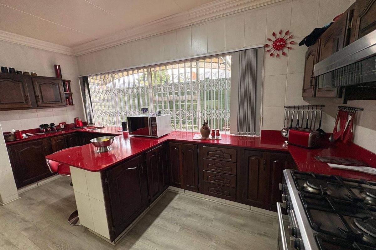 A functional kitchen featuring dark wood cabinetry, vibrant red countertops, a gas hob with extractor, and a large window with security bars.