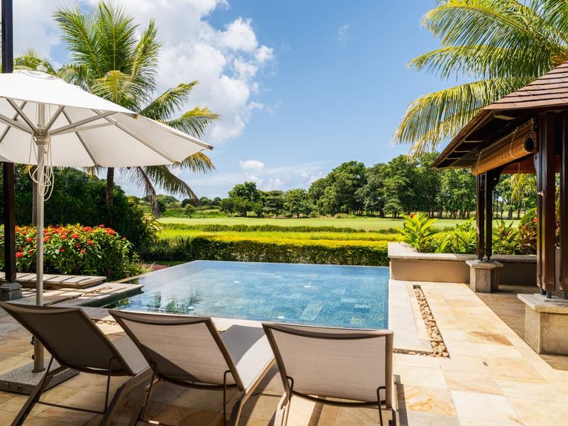 A serene outdoor pool area featuring a modern infinity-edge pool, stone-tiled patio, and a covered gazebo, overlooking a lush green landscape.