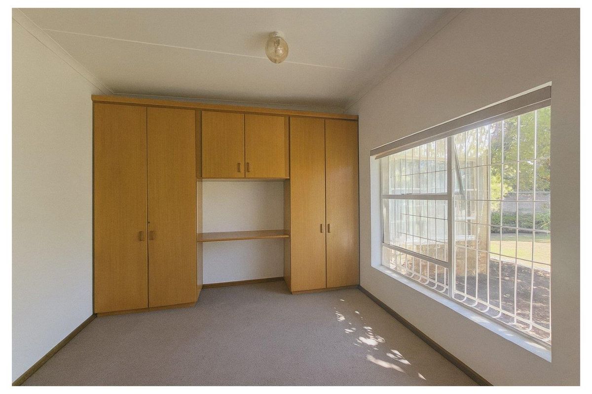 A bedroom featuring built-in wooden cabinetry with an integrated desk space and a large window fitted with security bars.