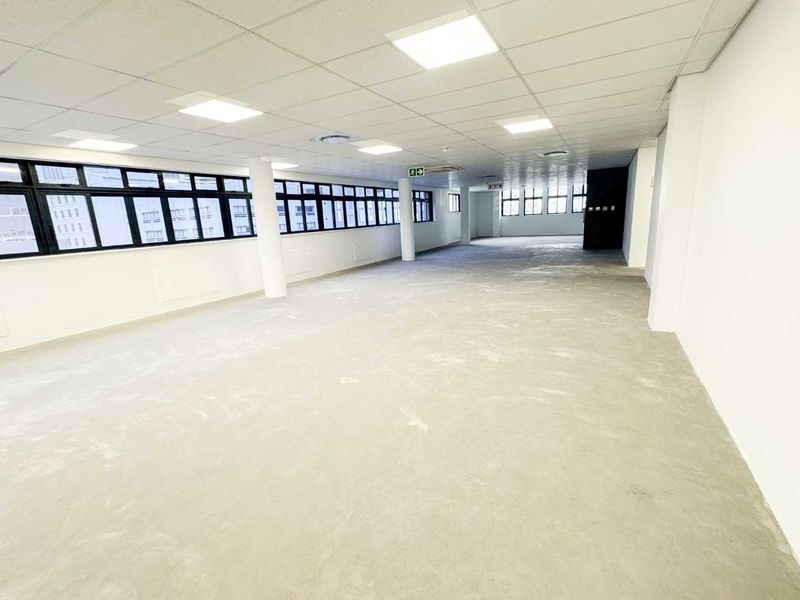 A spacious, open-plan office area featuring a suspended ceiling with recessed lighting, large windows providing natural light, and an unfinished concrete floor.