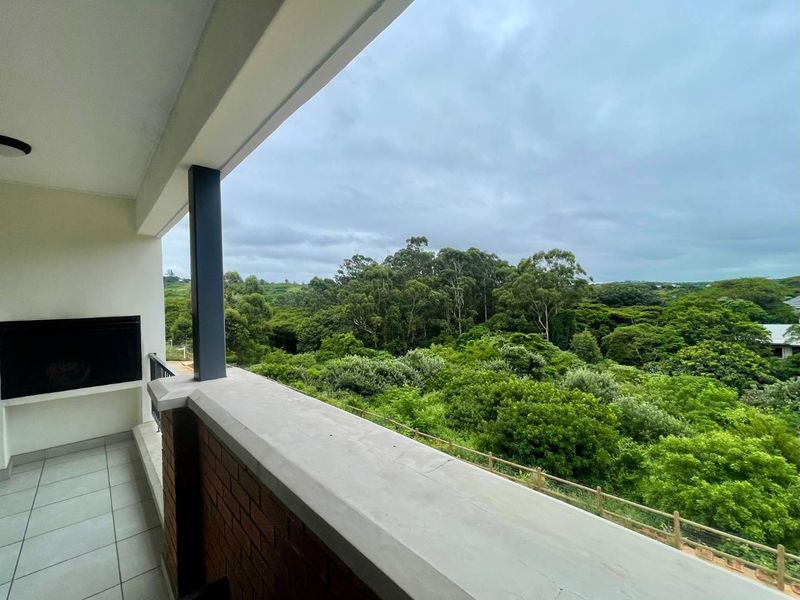 A covered balcony featuring a built-in braai, tiled flooring, and a brick balustrade, offering an elevated view of the surrounding lush greenery.